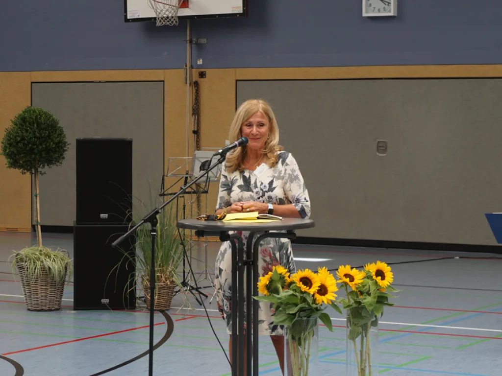 Person spricht am Rednerpult in der Sporthalle, davor Sonnenblumen.